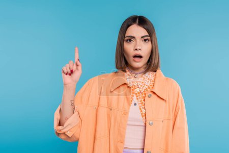 summer fashion, shocked young woman with short hair and piercing in nose and tattoos posing in casual outfit on blue background, everyday makeup, orange shirt, generation z, showing and pointing up