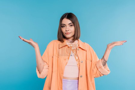 summer outfit, brunette young woman with short hair and piercing in nose and tattoos posing in casual outfit on blue background, orange shirt, generation z, not knowing and gesturing with hands