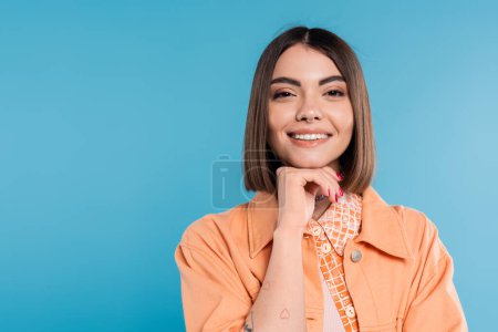 Sommermode, fröhliche junge Frau mit kurzen Haaren und Piercing in der Nase und Tätowierungen vor der Kamera auf blauem Hintergrund, Alltagsschminke, orangefarbenes Hemd, Generation z 