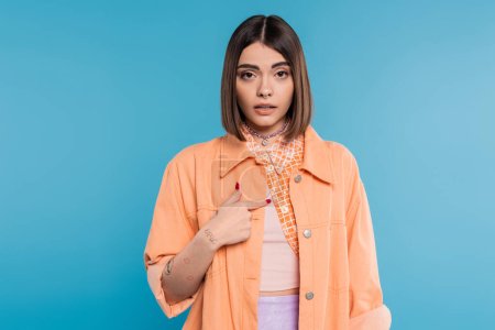 offended young woman pointing at herself with finger on blue background, looking at camera, summer outfit, emotional, displeased, generation z, short brunette hair, pierced nose, tattooed 