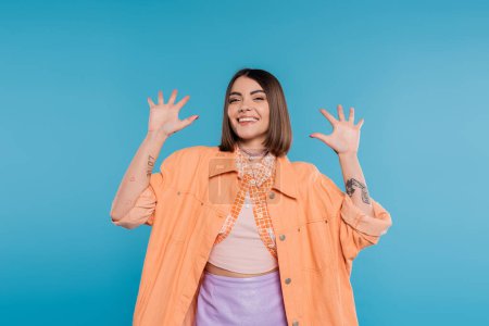 excited young woman gesturing and smiling while looking at camera, on blue background, summer outfit, generation z, short brunette hair, orange shirt, pierced nose, tattooed 