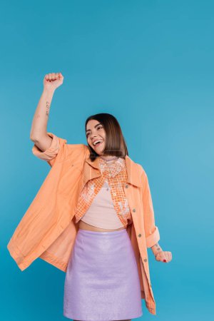 summer outfit, excited young woman smiling while gesturing on blue background, summer outfit, generation z, short brunette hair, orange shirt, pierced nose, tattooed 