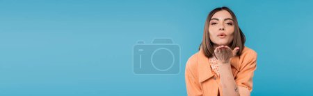 Photo for Sending air kiss, brunette woman with short hair, piercing in nose and tattoos posing in casual outfit on blue background, everyday makeup, orange shirt, generation z, blow kiss, banner - Royalty Free Image