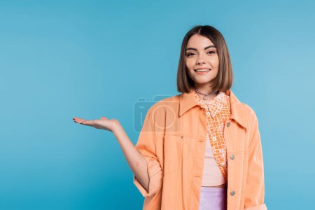 glückliche junge Frau mit kurzen Haaren, Piercing in der Nase und Tätowierungen, die etwas in der Kamera auf blauem Hintergrund vorführt, vorführend, orangefarbenes Hemd, Sommeroutfit Generation z 