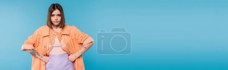 Photo for Offended woman standing with hands on hips on blue background, looking at camera, emotional, displeased, generation z, short brunette hair, pierced nose, tattooed, banner - Royalty Free Image