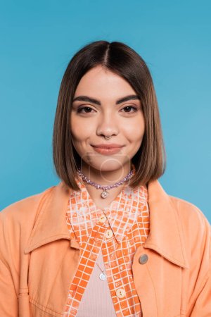 portrait of cheerful woman, young fashion model smiling and looking at camera on blue background, orange shirt, generation z, short brunette hair, pierced nose, summer outfit, gen z fashion 