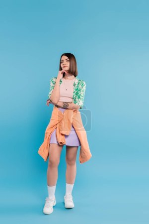generation z, young brunette woman with short hair posing in shirt with palm tree print, skirt and white sneakers on blue background, tattooed, nose piercing, casual attire, full length 