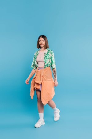 everyday style, young brunette woman with short hair walking in shirt with palm tree print, skirt and white sneakers on blue background, carefree, tattooed, nose piercing, casual attire, full length 