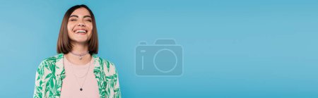 cheerful young woman with short brunette hair wearing shirt with palm tree print, smiling with closed eyes on blue background, casual attire, gen z fashion, emotional, happiness, banner 