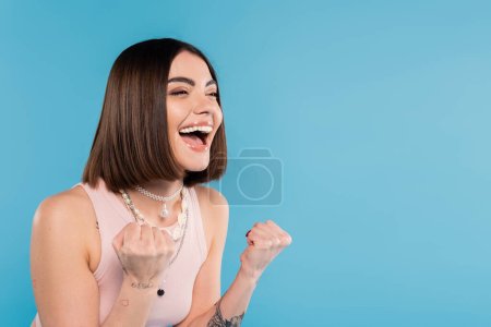 excitement, tattooed young woman with short brunette hair in tank top smiling and gesturing with hands on blue background, casual attire, gen z fashion, happiness 
