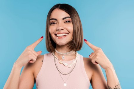excitement, tattooed young woman with short brunette hair in tank top smiling and pointing at her cheeks on blue background, casual attire, gen z fashion, happiness, joyful 