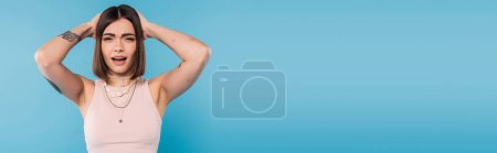 Photo for Failure, frustrated, young woman with short brunette hair and tattoos holding hands near head and looking at camera on blue background, casual attire, gen z fashion, emotional, banner - Royalty Free Image