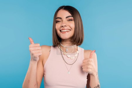 positivity, tattooed young woman with short brunette hair in tank top smiling and showing thumbs up on blue background, casual attire, gen z fashion, happiness, like gesture