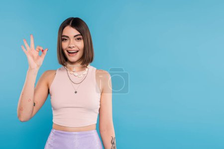 excitement, tattooed young woman with short hair in tank top smiling and showing ok gesture on blue background, casual attire, gen z fashion, fashionable trend, happiness 