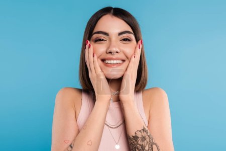 positivity, tattooed young woman with short brunette hair in tank top smiling  and touching her face on blue background, casual attire, gen z fashion, happiness, joyful face