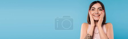 Foto de Positividad, mujer joven tatuada con pelo corto en camiseta sin mangas sonriendo y tocando su cara sobre fondo azul, atuendo casual, moda gen z, felicidad, cara alegre, pancarta - Imagen libre de derechos