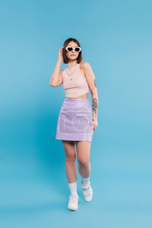 fashion trend, brunette young woman with short hair in tank top, skirt, white sneakers and sunglasses posing on blue background, casual attire, gen z fashion, personal style, nose piercing 