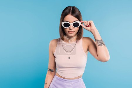 gen z fashion, brunette young woman with short hair in tank top, skirt and sunglasses posing on blue background, casual attire, stylish posing, personal style, portrait, fashionista 