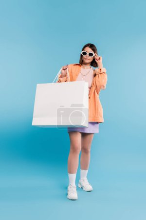 shopping spree, jeune femme brune en débardeur, jupe, lunettes de soleil et chemise orange posant avec sac à provisions sur fond bleu, tenue décontractée, pose stylée, gen z, mode moderne 