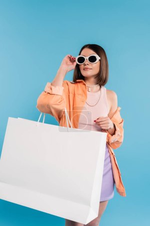shopping d'été, jeune femme à la mode en débardeur, jupe, lunettes de soleil et chemise orange posant avec sac à provisions sur fond bleu, tenue décontractée, pose élégante, gen z, mode moderne 