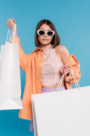 Sommereinkauf, Generation Z, brünette junge Frau im Tank-Top, Rock, Sonnenbrille und orangefarbenem Hemd posiert mit Einkaufstaschen auf blauem Hintergrund, lässige Kleidung, stilvolles Posen, moderne Mode 