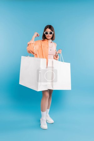 shopping d'été, tendance de la mode, jeune femme brune en débardeur, jupe, lunettes de soleil et chemise orange posant avec des sacs à provisions sur fond bleu, pose élégante, gen z, mode moderne, pleine longueur 
