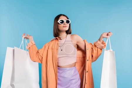 Sommerspaß, brünette junge Frau mit Sonnenbrille und trendigem Outfit posiert mit Einkaufstaschen auf blauem Hintergrund, lässige Kleidung, stilvolles Posing, Generation Z, moderne Mode 
