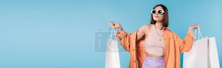 summer spree, brunette young woman in sunglasses and trendy outfit posing with shopping bags on blue background, casual attire, stylish posing, generation z, fashion trend, banner 