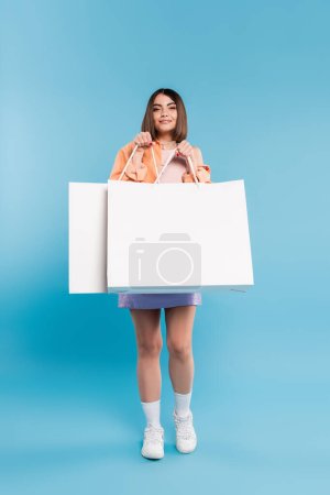 Positivität, Einkaufsbummel, fröhliche junge Frau im trendigen Outfit posiert mit Einkaufstaschen auf blauem Hintergrund, lässige Kleidung, stilvoll, Generation Z, moderne Mode 