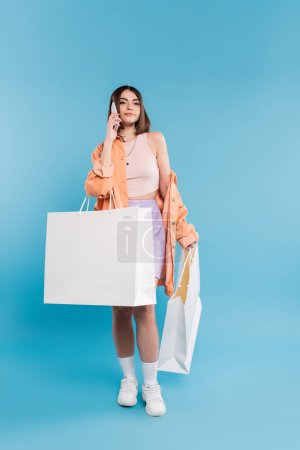 positivity, shopping spree, phone call, cheerful young woman in trendy outfit holding shopping bags and talking on smartphone on blue background, casual attire, generation z, modern fashion 
