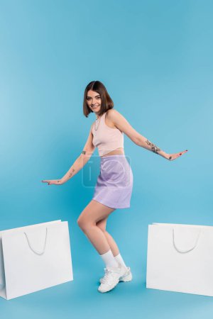 positivity, summer fashion, shopping spree, tattooed young woman with nose piercing posing in trendy outfit near shopping bags on blue background, casual attire, generation z, modern fashion 
