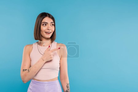 Photo for Amazed woman showing something and looking away, stylish and tattooed model with nose piercing pointing with finger on blue background, generation z, summer fashion, smile - Royalty Free Image