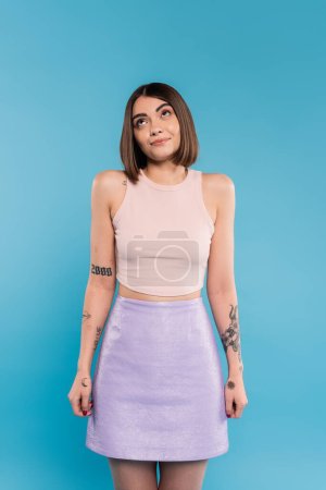 not knowing, smiling young woman with tattoos and nose piercing standing in tank top and skirt on blue background, looking up, confused, pretty face, generation z, summer outfit 