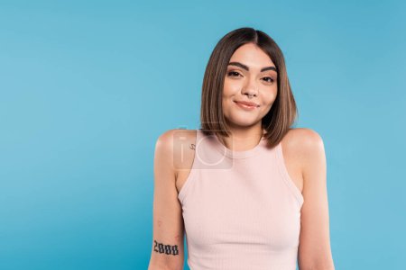 not knowing, smiling young woman with tattoos and nose piercing standing in tank top on blue background, looking at camera, confused, pretty face, generation z, summer outfit 