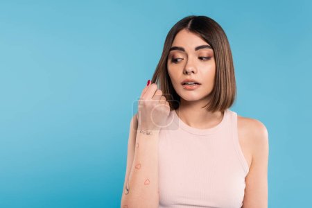 hair damage, stress, concerned brunette young woman touching her hair on blue background, tattooed, nose piercing, tank top, generation z, casual attire, thinning hair 