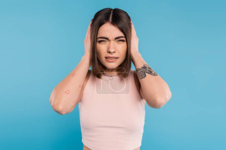 migraine, tattooed young woman with nose piercing and short hair touching head while suffering from headache on blue background, generation z, tiredness, stress 
