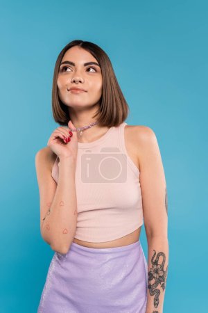dreamy, tattooed young woman with nose piercing and short hair touching silver necklace and looking away on blue background, generation z, fashionable, trendy summer fashion 