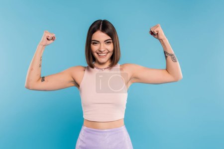 women power, cheerful young woman with short hair, tattoos and nose piercing showing muscles on blue background, generation z, displeased, casual attire, strength 