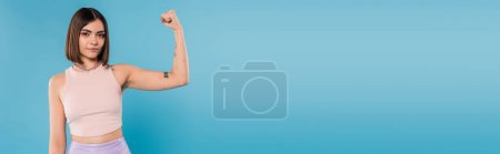 female power, brunette young woman with short hair, tattoos and nose piercing showing muscle on blue background, generation z, displeased, casual attire, strength, banner 