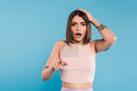shocked face, brunette young woman with short hair, tattoos and nose piercing pointing at camera on blue background, generation z, displeased, casual attire, summer outfit 