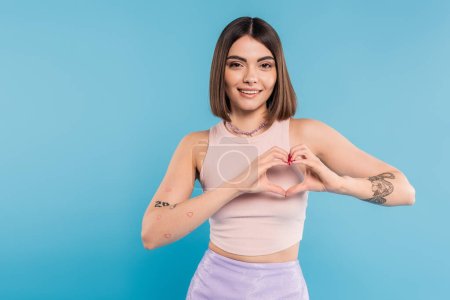 positivity and love, happy young woman with short hair, tattoos and nose piercing showing heart gesture with hands on blue background, generation z, cheerful, casual attire