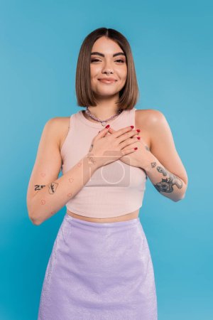 positivity and peace, happy young woman with short hair, tattoos and nose piercing holding hands near chest on blue background, generation z, cheerful, casual attire 