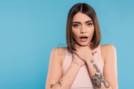 brunette young woman with short hair holding hands near neck and standing with opened mouth while choking on blue background, casual attire, gen z fashion, personal style, nose piercing 