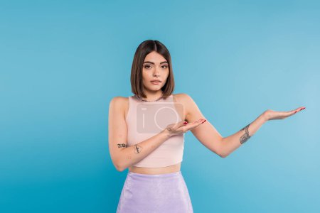 brunette young woman with short hair demonstrating something on camera on blue background, casual attire, gen z fashion, personal style, nose piercing, pointing with hands 