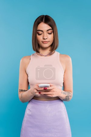 Foto de Mujer mensajes de texto en el teléfono inteligente, pelo corto, tatuajes y piercing en la nariz usando el teléfono móvil sobre fondo azul, atuendo casual, moda gen z, estilo personal, maquillaje diario - Imagen libre de derechos