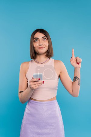 Photo for Holding smartphone, young brunette woman short hair, tattoos and nose piercing pointing up on blue background, casual attire, gen z fashion, social media influencers - Royalty Free Image