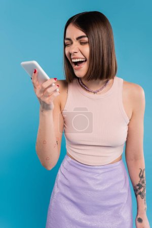 Botschaften senden, aufgeregte junge Frau kurze Haare, Tätowierungen und Nasenpiercing mit dem Handy auf blauem Hintergrund, lässige Kleidung, Gen-Z-Mode, Social-Media-Influencer 