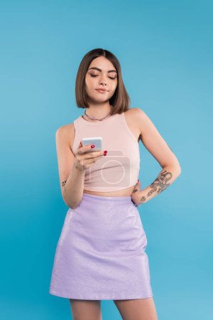 Senden einer Nachricht, attraktive junge Frau kurze Haare, Tätowierungen und Nasenpiercing mit dem Handy auf blauem Hintergrund, lässige Kleidung, Gen-Z-Mode, Social-Media-Influencer 