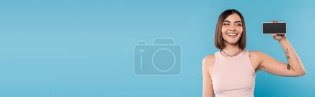 Photo for Smartphone with blank screen, attractive young woman with short hair, tattoos and nose piercing holding mobile phone on blue background, gen z fashion, social media influencers, banner - Royalty Free Image
