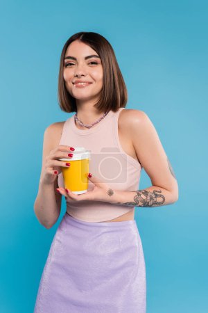 to go coffee, happy young woman with short hair, tattoos and nose piercing holding paper cup on blue background, generation z, summer trends, attractive, coffee culture, everyday style 